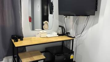 A compact room with a desk, a chair, a small black storage unit, a television mounted on the wall, and a mirror reflecting the room.