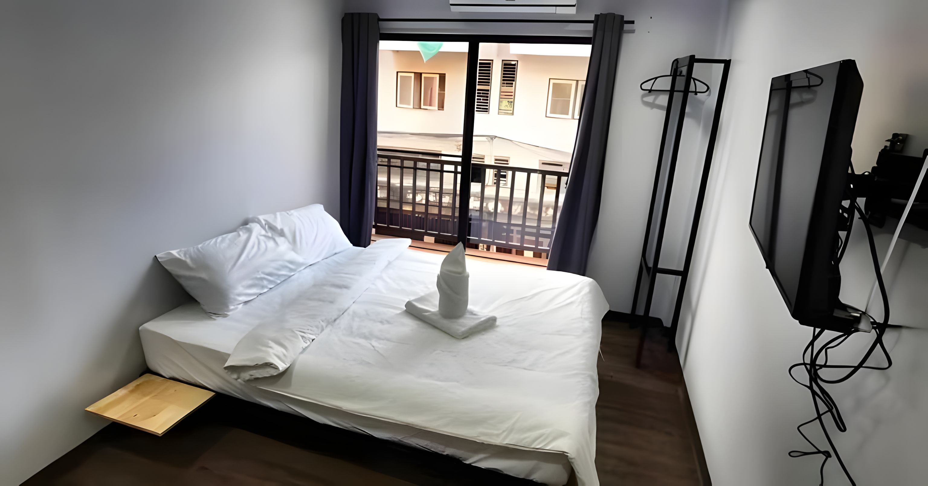 A hotel room with a bed, a small wooden side table, a balcony with a railing, and a television mounted on the wall.