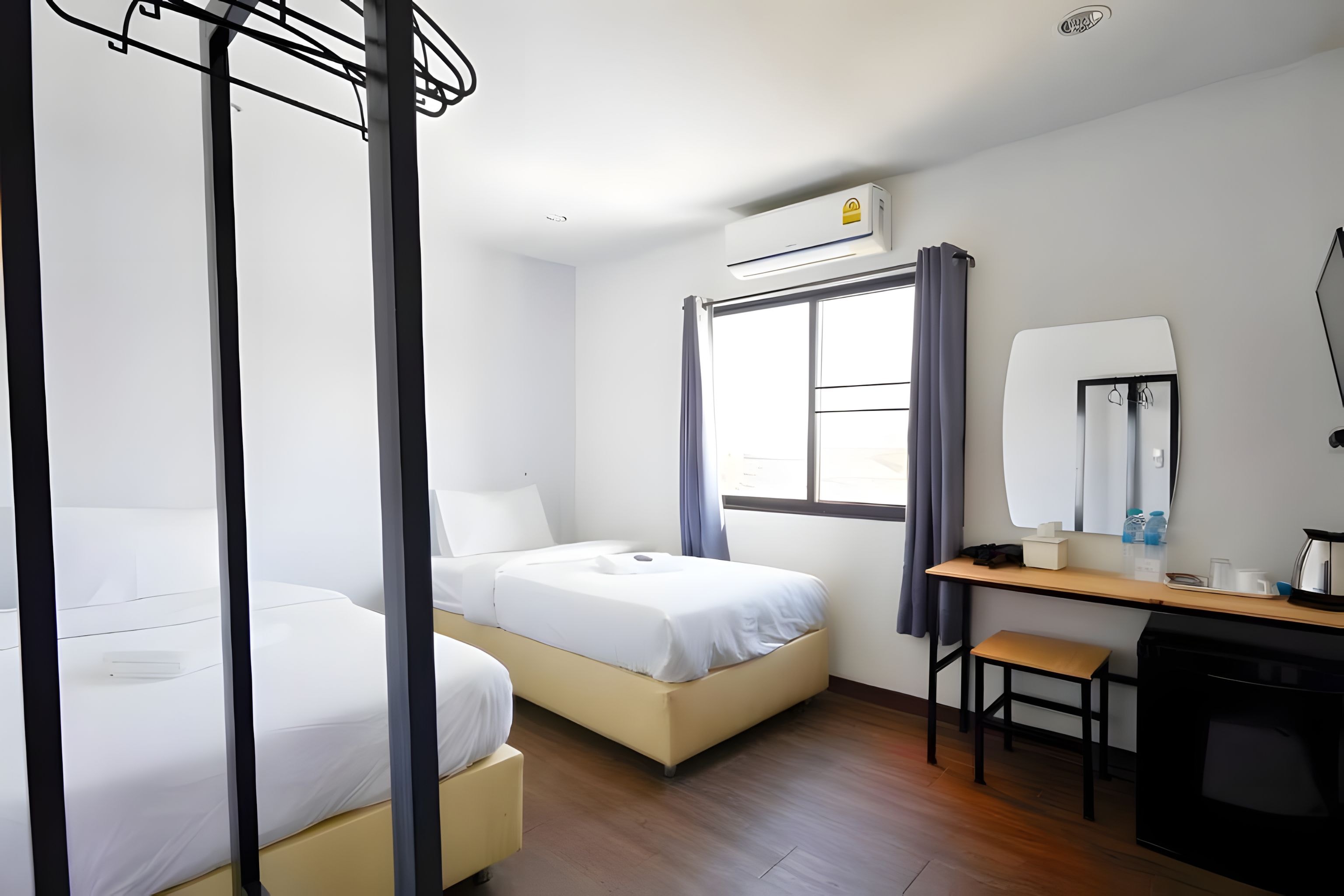 A hotel room with two single beds, a desk with a chair, a window with curtains, and a small kitchenette area.