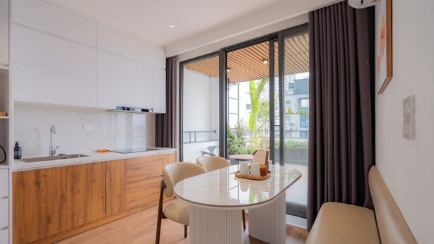 A modern kitchen with white cabinets, a wooden countertop, and a dining area with a round table and chairs. - Mia Apartments by XÔI Residences (Hanoi)