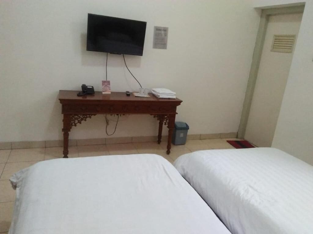 A room with two beds, a wooden desk with a telephone and a small item on it, a television mounted on the wall, and a trash bin.