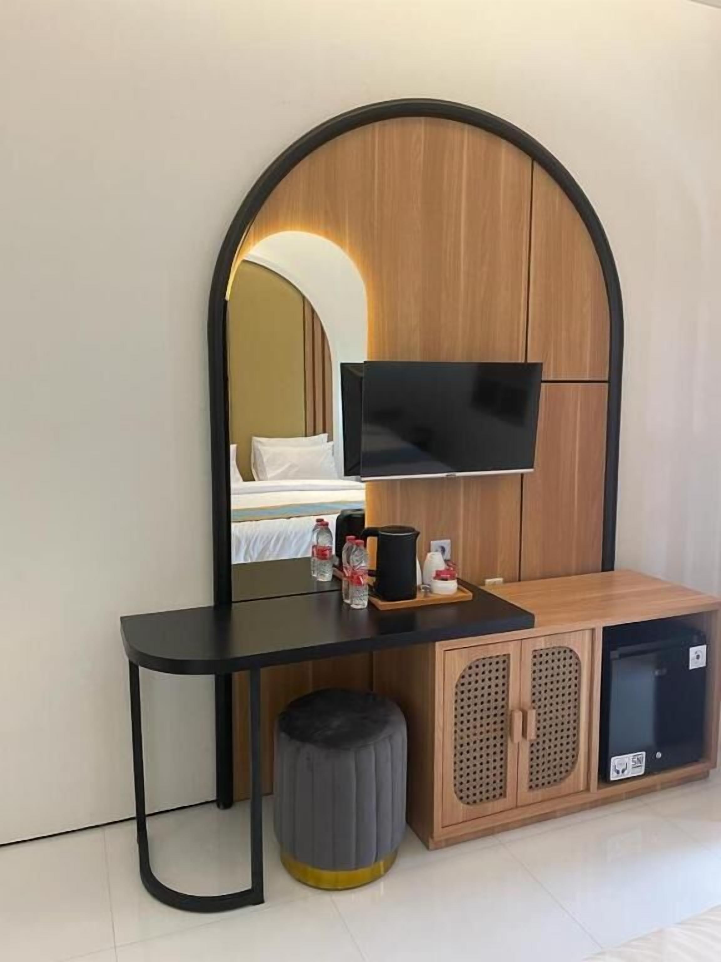 Une chambre d’hôtel moderne avec une coiffeuse en bois, un bureau noir, un téléviseur fixé sur un panneau en bois, un petit tabouret et un meuble contenant un réfrigérateur.