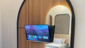 Une chambre d’hôtel dotée d’un téléviseur à écran plat fixé sur un mur en bois, d’un lit avec une tête de lit blanche, d’une table de chevet sur laquelle se trouve un plateau avec des articles de toilette, et d’une petite poubelle.