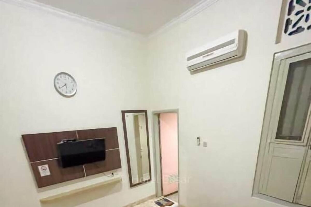 A hotel room with a bed, a TV mounted on the wall, a clock, and an air conditioning unit.