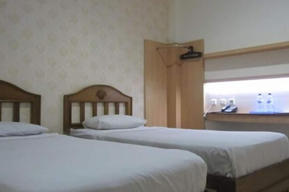 A room with two single beds, a wooden headboard, a desk with a chair, and a window with a light fixture.