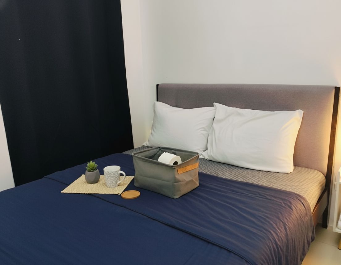 Una cama bien hecha con una colcha azul oscuro, dos almohadas blancas, una caja gris con un rollo de papel higiénico, una pequeña planta en maceta y una taza blanca sobre un posavasos tejido.