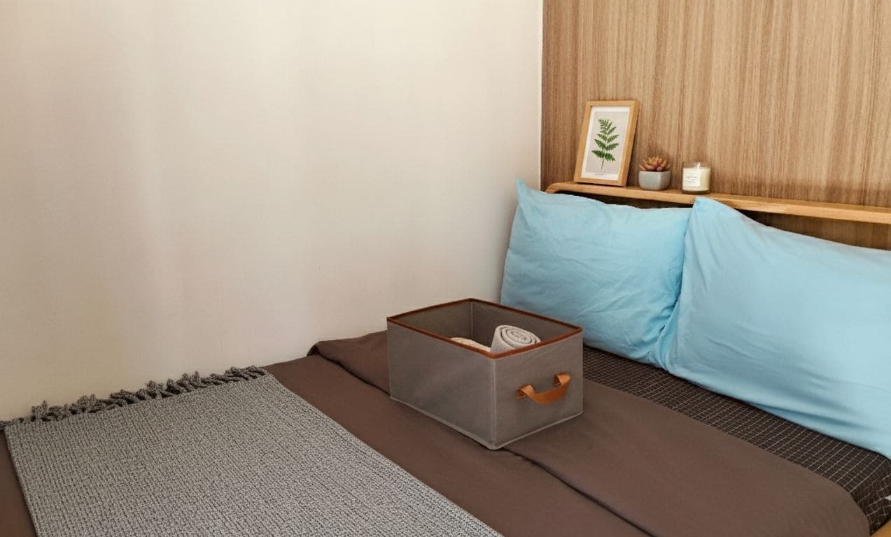 A neatly arranged bedroom with a bed, a grey storage box, a blue pillow, and a wooden shelf with a plant and candle.