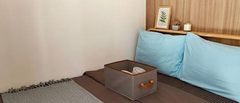 A neatly arranged bedroom with a bed, a grey storage box, a blue pillow, and a wooden shelf with a plant and candle.
