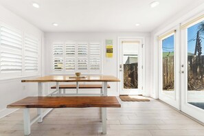 A dining area with a wooden table and benches, white walls, and a view through glass doors.