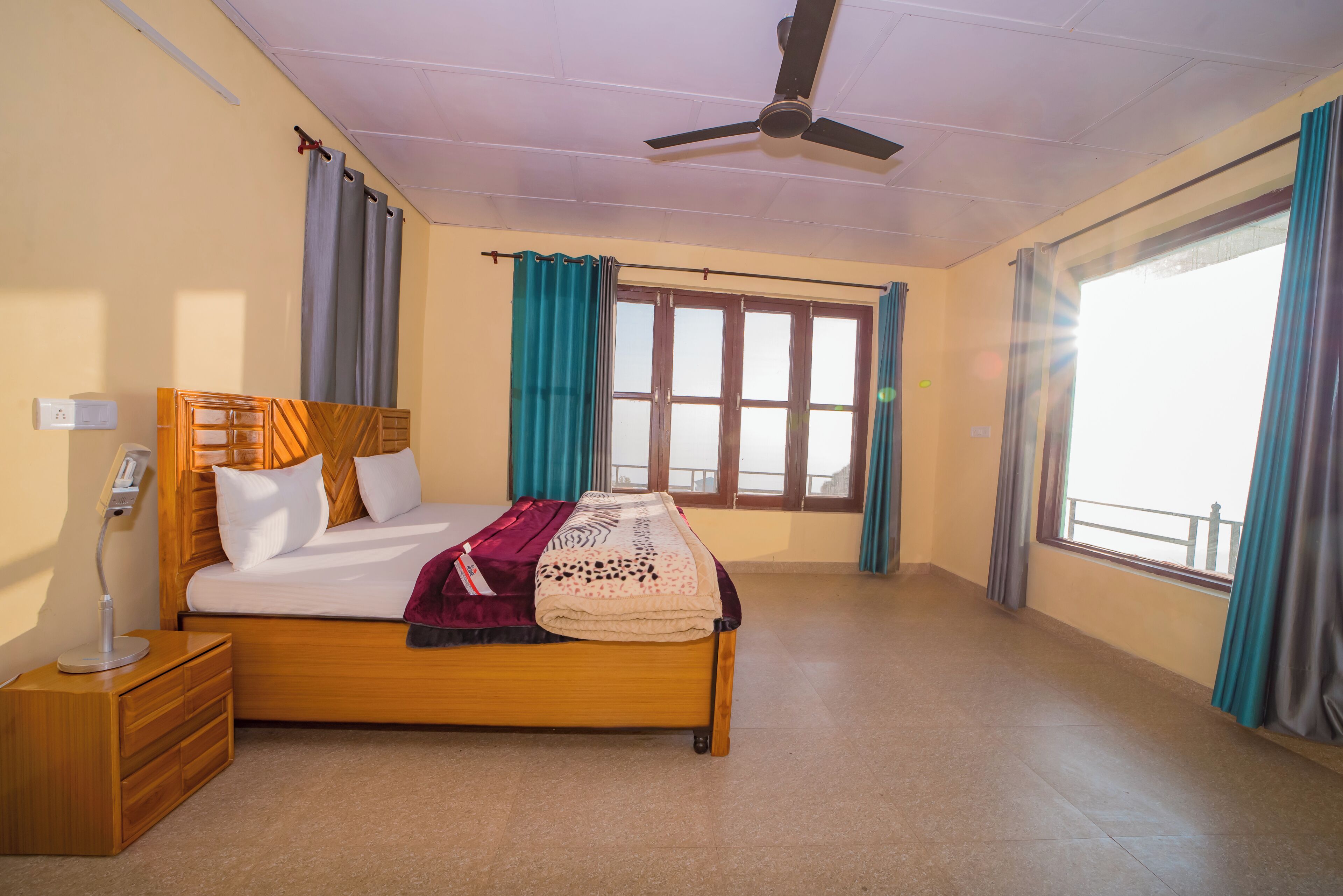 Un dormitorio con cama, mesita de noche, lámpara de noche, ventilador de techo, ventanas con cortinas y vistas al exterior.