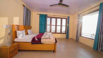 Un dormitorio con cama, mesita de noche, lámpara de noche, ventilador de techo, ventanas con cortinas y vistas al exterior.