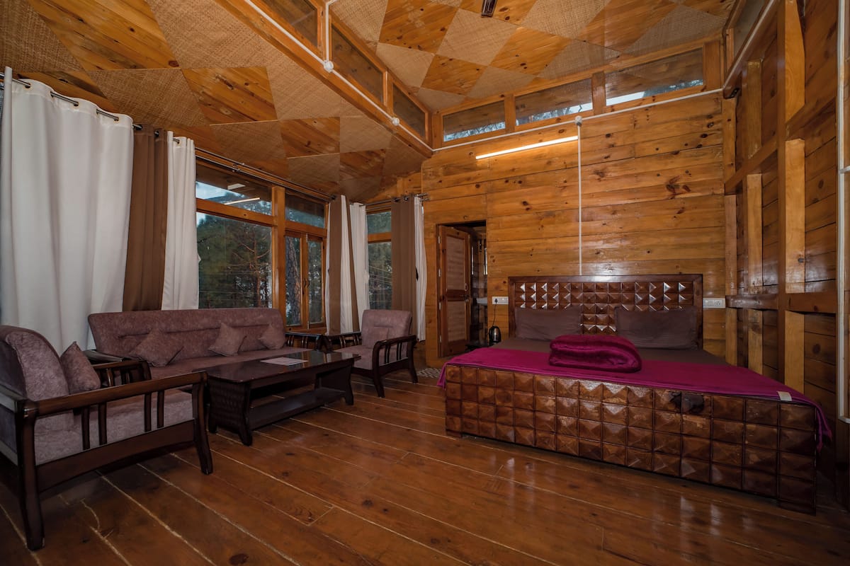 A wooden cabin interior with a sofa, chairs, a bed, and large windows.