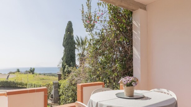 Un balcon avec une table et des chaises, donnant sur un paysage pittoresque, avec des arbres et une vue sur la mer.