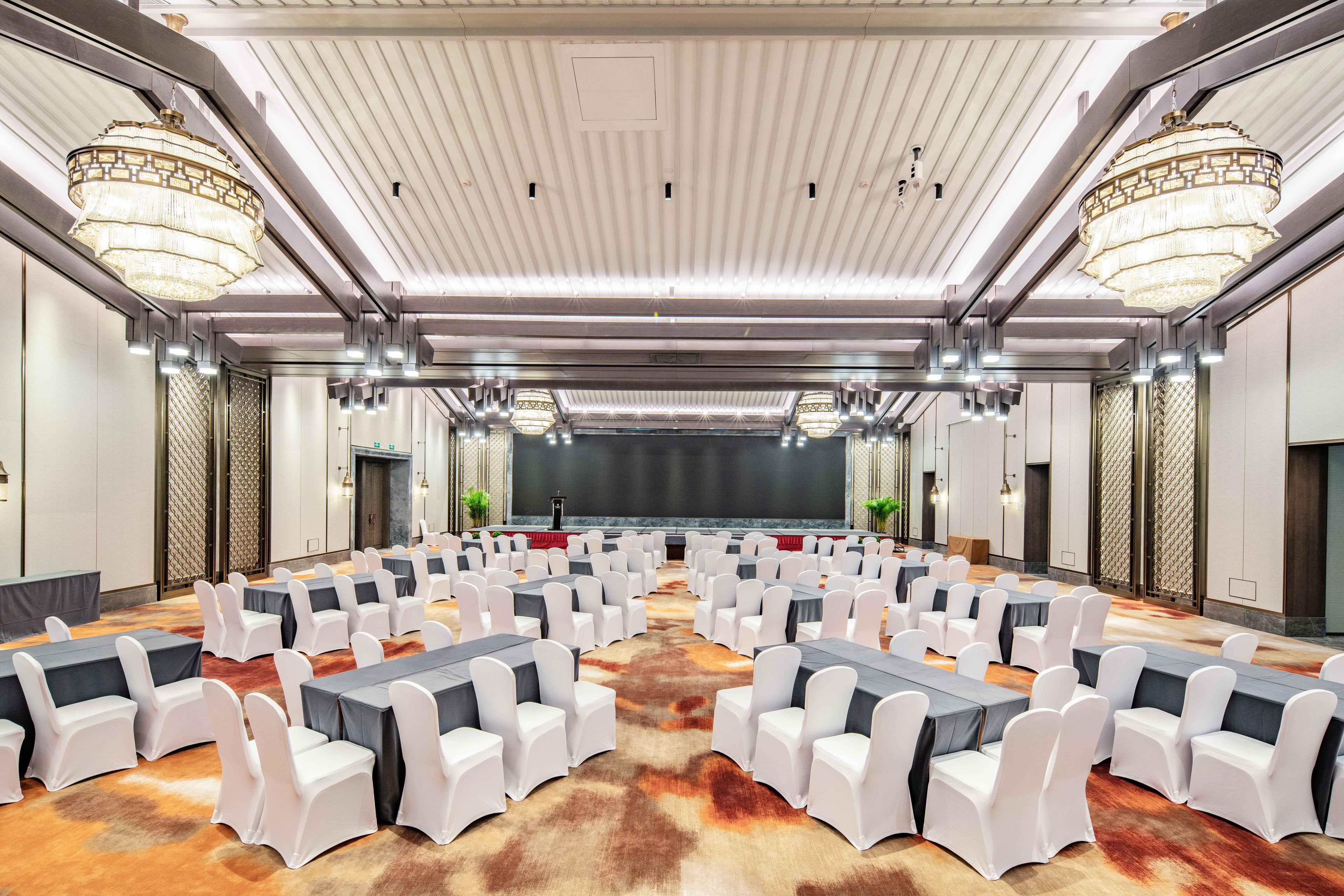 Un salón de conferencias con filas de mesas y sillas, un escenario y candelabros.