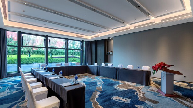 A conference room with a long table, white chairs, a podium, and a large window with a view of greenery. - Mauve Glamor Chengdu (Chengdu)