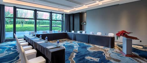 A conference room with a long table, white chairs, a podium, and a large window with a view of greenery.