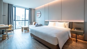 A modern hotel room with a large bed, a desk, a sofa, and a view of the city through large windows. - Mauve Glamor Chengdu (Chengdu)