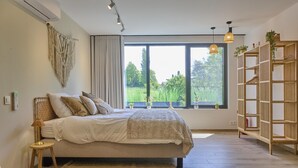 A bedroom with a large bed, a bedside table, a lamp, a window with a view of greenery, and a wooden shelf with plants.