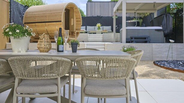 An outdoor patio with a table set for two, surrounded by wicker chairs, a wooden barrel, and potted plants.