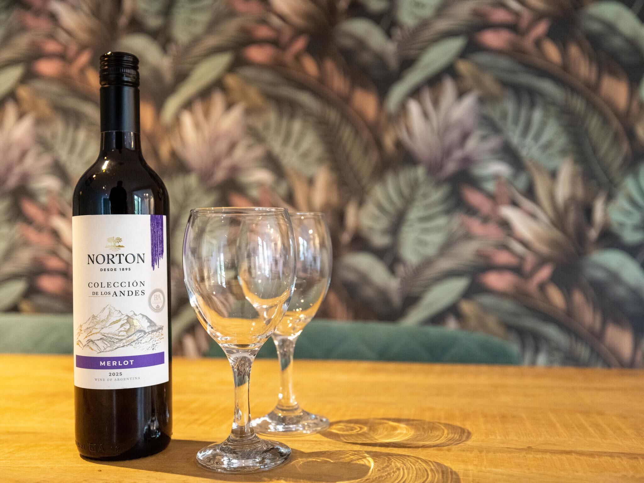 A bottle of Norton wine and two glasses on a wooden table with a patterned background.