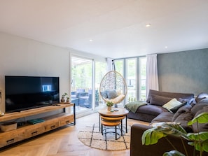 A modern living room with a flat-screen TV, a hanging chair, a sofa, and a dining table. - Charming chalet on a campsite with outdoor pool (IJhorst)