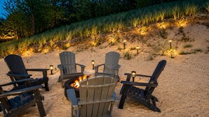 Uma fogueira cercada por quatro cadeiras Adirondack, com lanternas, situada numa praia arenosa com grama e árvores ao fundo.