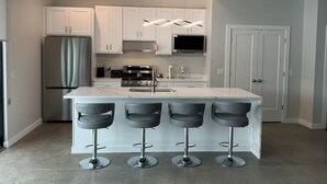 Uma cozinha moderna com armários brancos, uma geladeira de aço inoxidável, uma ilha central com banquetas e iluminação embutida no teto.