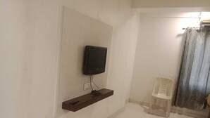 Una stanza con una TV montata a parete, un piccolo ripiano con un telecomando, una sedia di plastica bianca e una tenda.