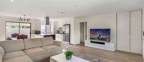 A modern living room with a sofa, a coffee table with a plant, a flat-screen TV, and a kitchen in the background.