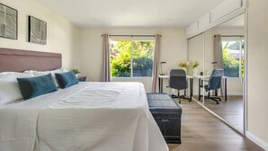 Ein Schlafzimmer mit einem großen Bett, einem Schreibtisch und einem Sessel. Ein Fenster mit Vorhängen bietet Blick auf viel Grün.