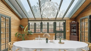 A wooden dining room with a table set for four, surrounded by chairs, a large window with a view of trees, and a cabinet with glassware. - Forest retreat with sauna & nearby beach (Højby)
