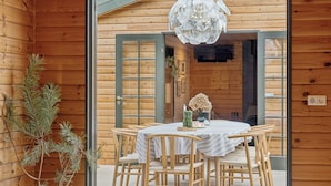 A wooden cabin interior with a dining table, chairs, a hanging light fixture, and a pine tree. - Forest retreat with sauna & nearby beach (Højby)