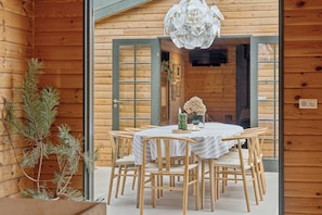 A wooden cabin interior with a dining table, chairs, a hanging light fixture, and a pine tree. - Forest retreat with sauna & nearby beach (Højby)