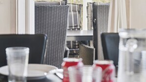 Uma área de refeições com cadeiras de vime, uma mesa com pratos e copos, e vista pela janela.