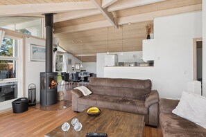 A modern living room with a wood-burning stove, a brown leather sofa, a dining area, and a kitchen in the background.