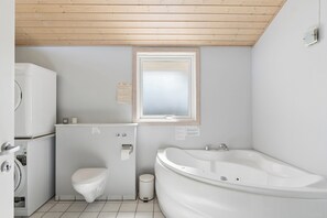 A compact bathroom with a toilet, washing machine, and bathtub. - Spa holiday home by Høve Beach (Asnæs)