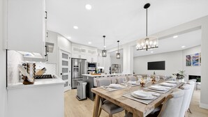 A modern kitchen with white cabinetry, a dining area with a wooden table set for six, and a large island with a built-in oven. - Modern Luxury 6BR Home •Sleeps12•12 mins Airport (Calgary)