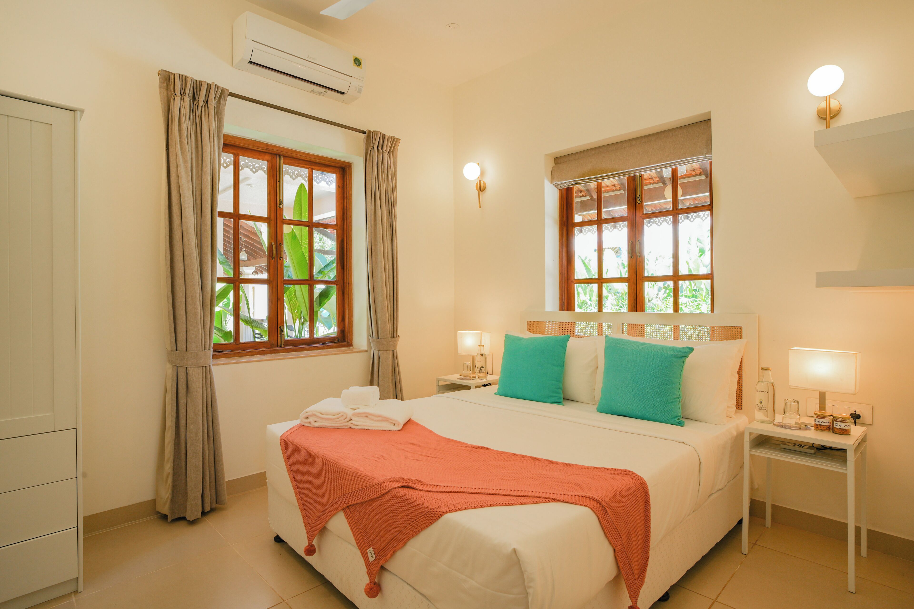 A hotel room with a bed, two bedside tables, a window with curtains, an air conditioning unit, and a wall-mounted light fixture.