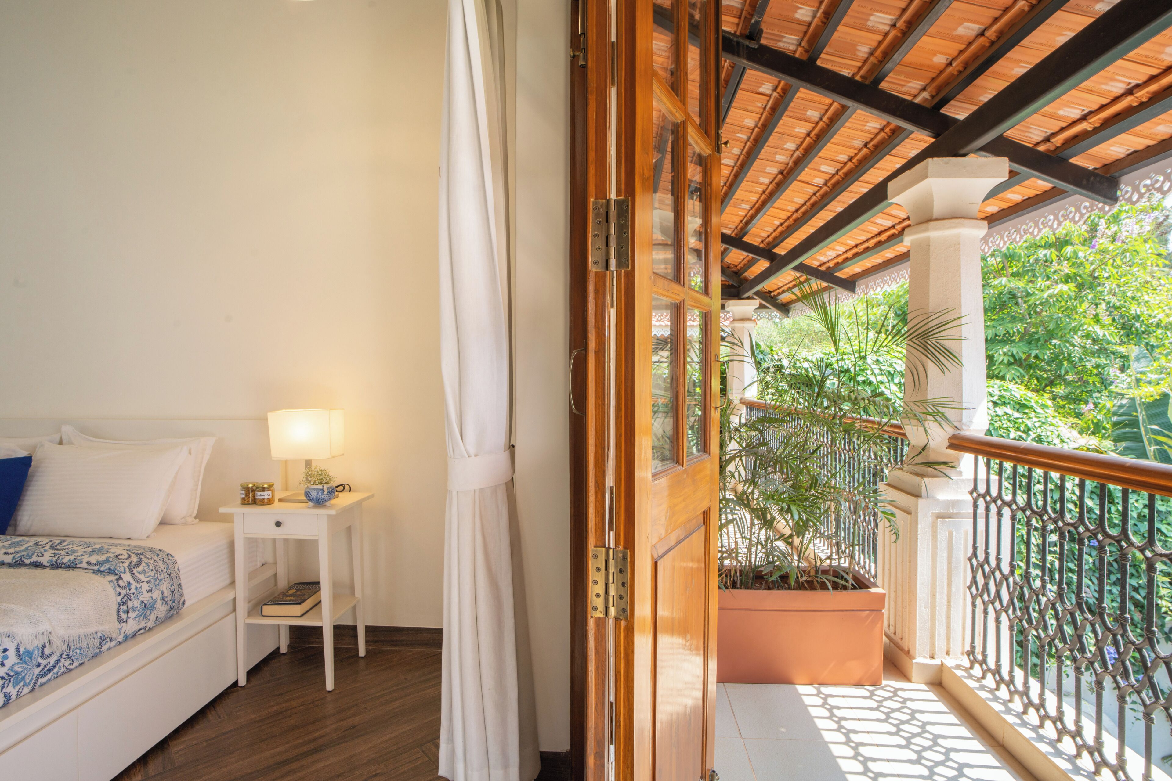 Un dormitorio con cama, mesita de noche, lámpara y vistas a un balcón con plantas.