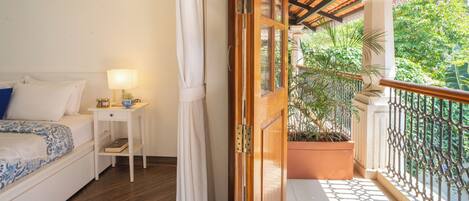 Un dormitorio con cama, mesita de noche, lámpara y vistas a un balcón con plantas.