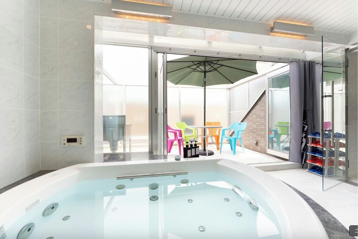 Ein modernes Badezimmer mit einer gläsernen Eckbadewanne und einer Glastür, die auf einen Balkon mit bunten Stühlen und einem Tisch führt.