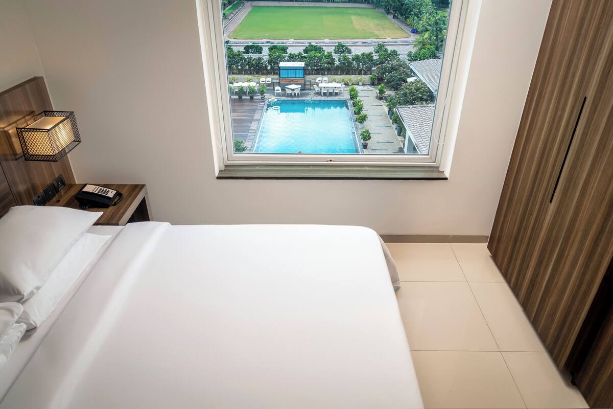 Una habitación de hotel con una cama, una mesita de noche, una lámpara y un ventanal con vista a una piscina y follaje.