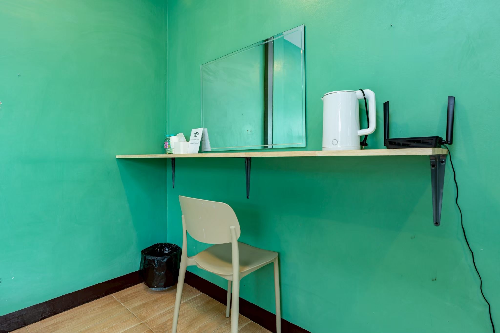A small room with teal walls, a wooden floor, a white chair, a trash bin, a shelf with a kettle, a mirror, and a black speaker.