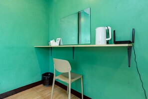 Een kleine kamer met turquoise wanden, een houten vloer, een witte stoel, een prullenbak, een plankje met een waterkoker, een spiegel en een zwarte speaker.