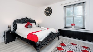 Un dormitorio con una cama negra ornamentada, ropa de cama blanca, un plaid rojo, un reloj de pared, una ventana con persianas y un radiador.