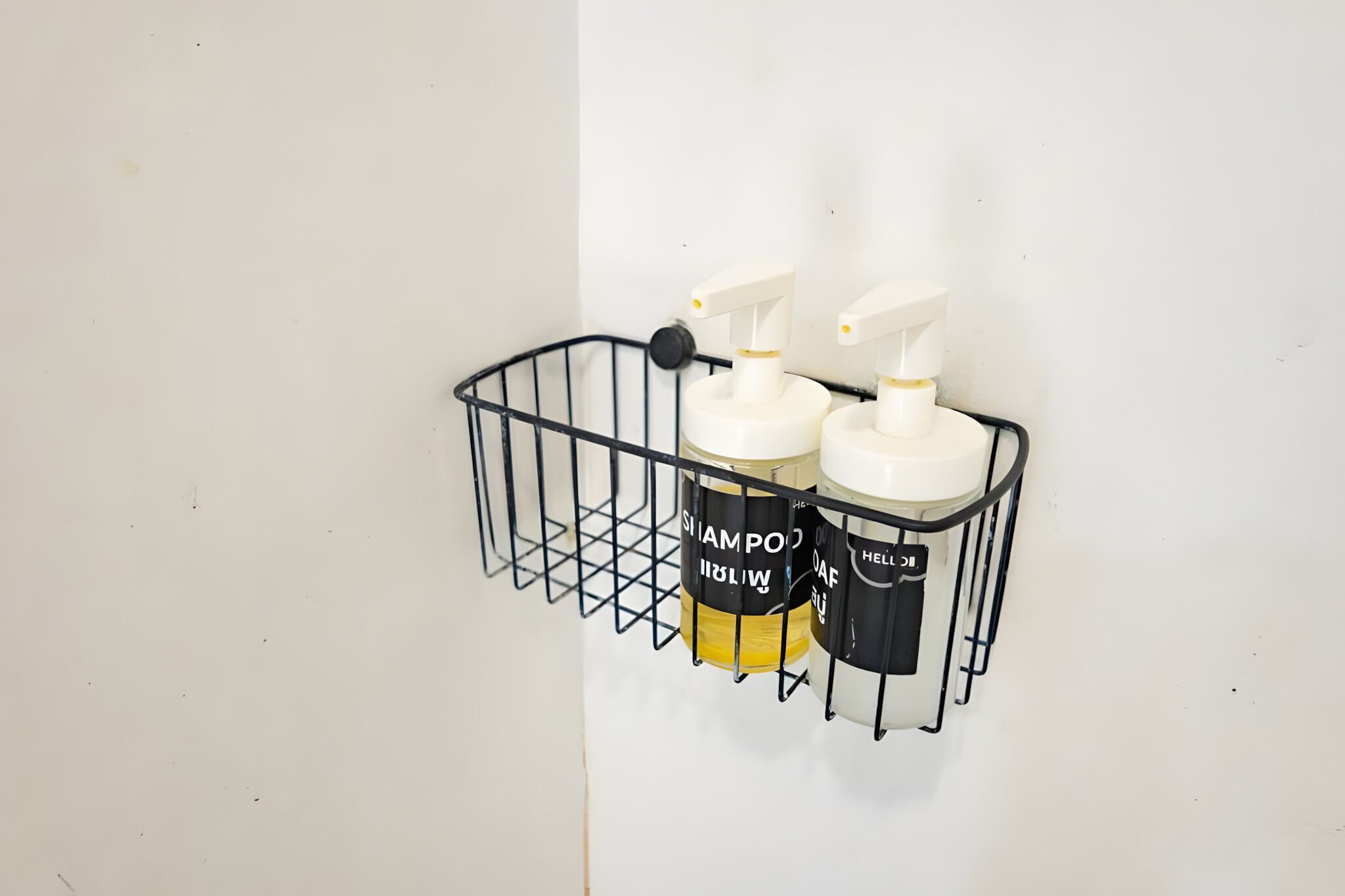 En sort metalkasse med to hvide og en gul shampoo-flaske, mærket 'SHAMPOO' og 'HELLO', monteret på en hvid væg.