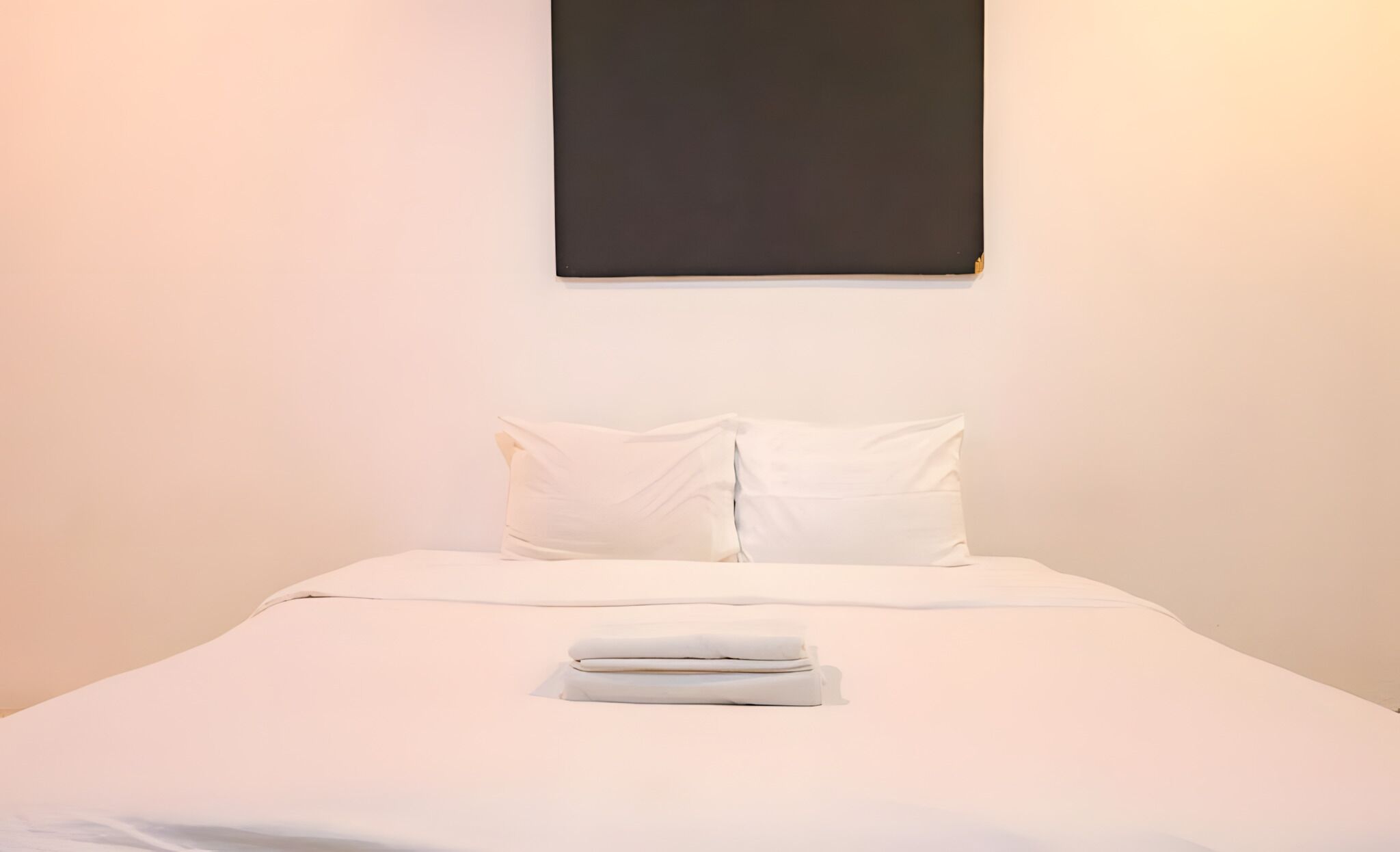 A neatly made bed with white linens and pillows, a folded towel, and a dark-colored artwork on the wall.