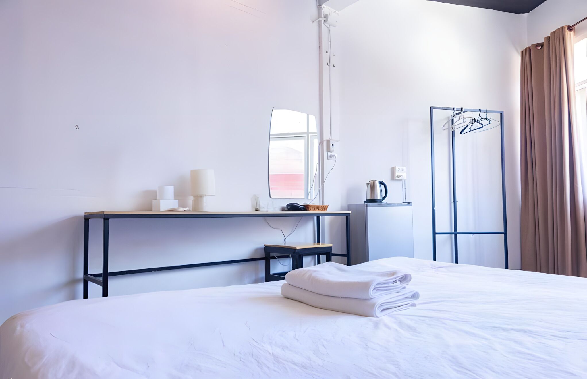 A hotel room with a bed, a desk with a lamp, a mirror, a radiator, and a window with curtains.