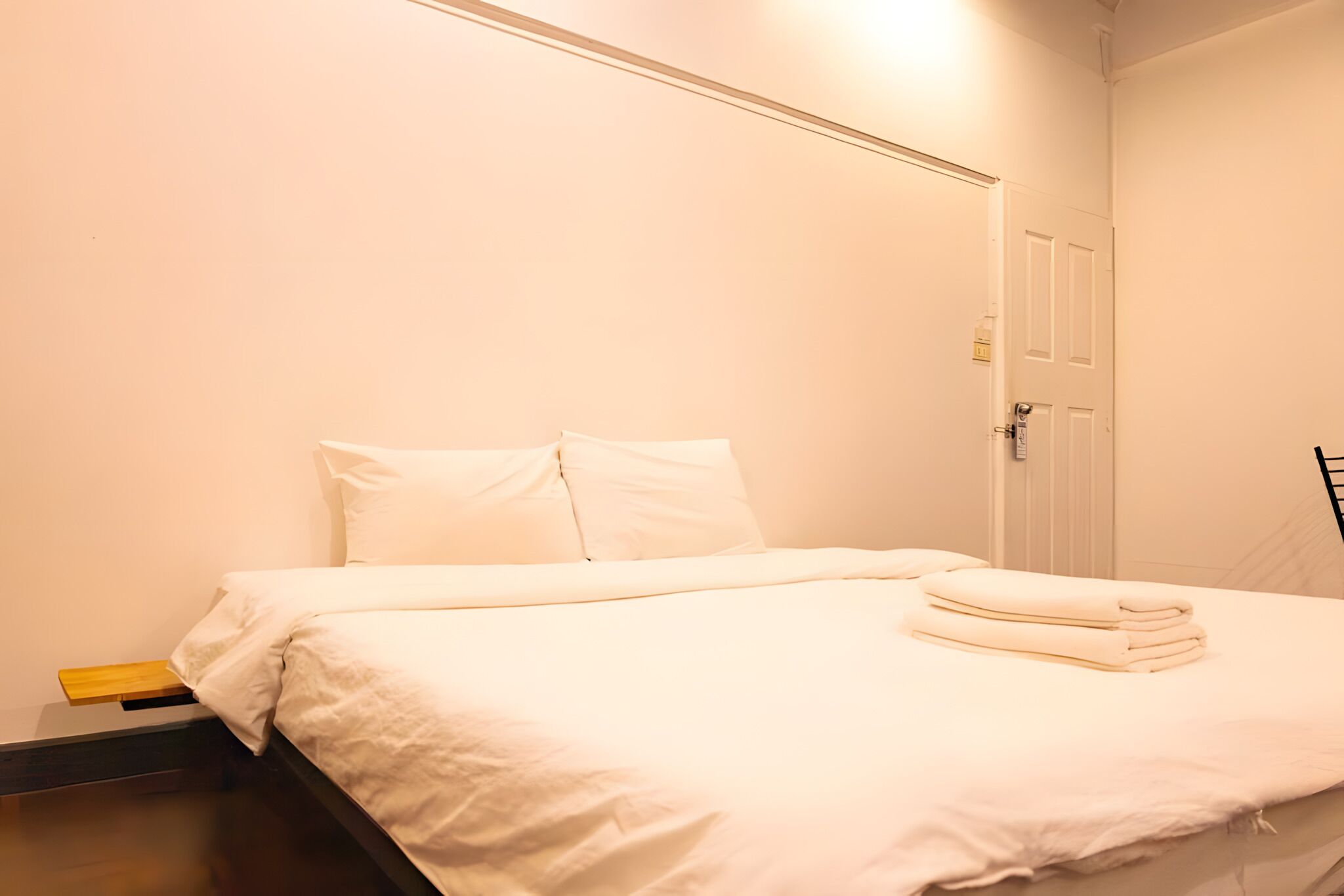 Una cama bien tendida con sábanas blancas, una mesita de noche de madera y una puerta con picaporte.