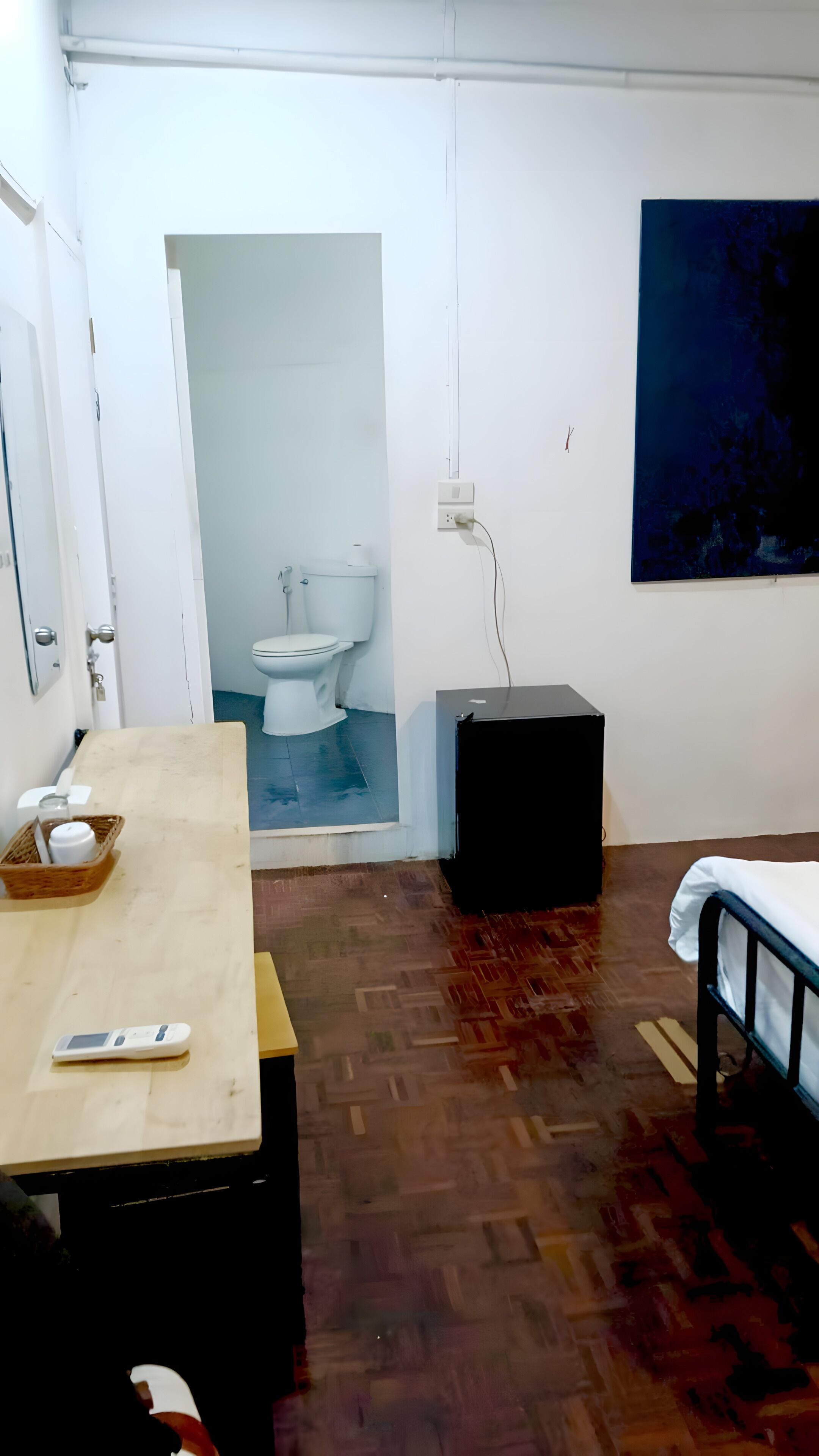 A room with a wooden floor, a bed, a desk with a mobile phone, a small table with a basket, a black speaker, and a painting on the wall.
